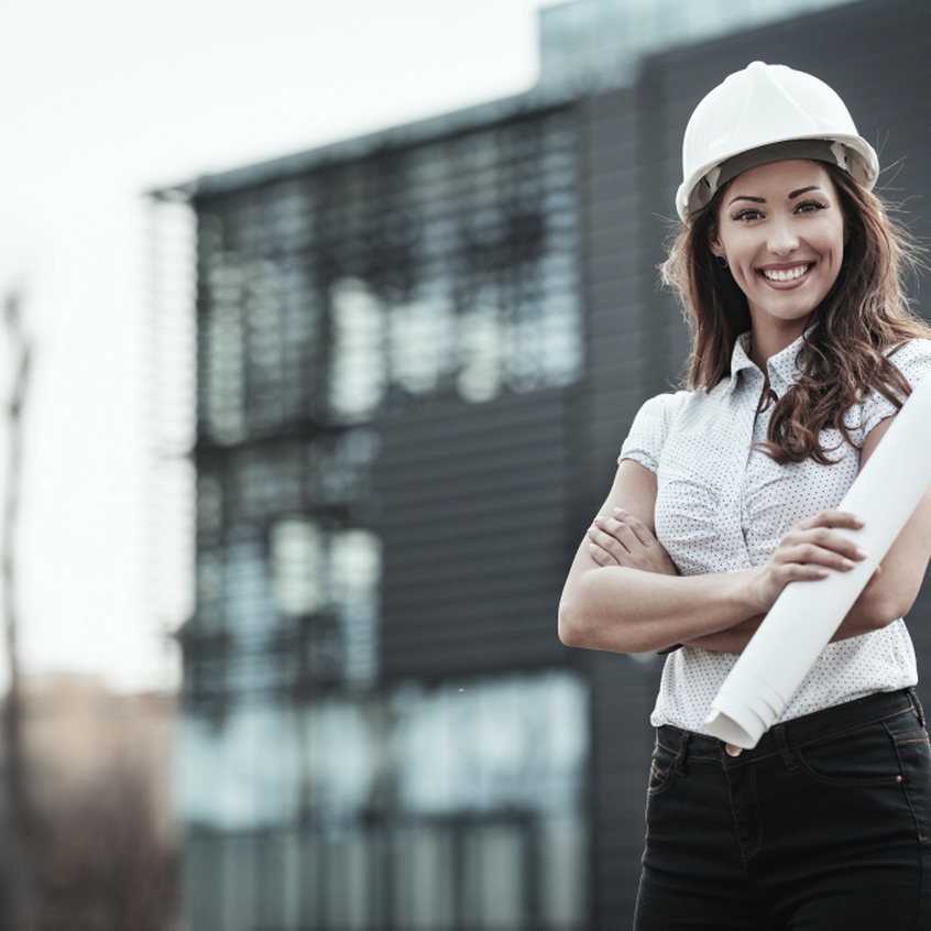 Engenheira de capacete branco, de braços cruzados, segurando um projeto impresso
